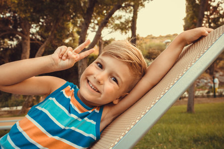 Smiling little boy showing victory sing toward the camera while relaxing on deck chair.の写真素材