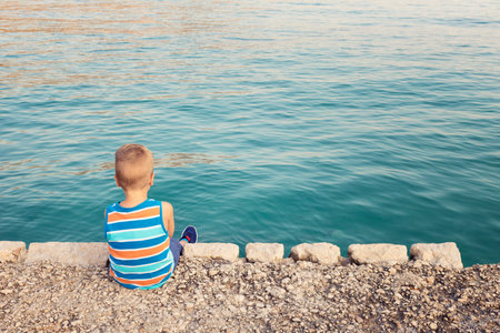 Back view of lonely child relaxing on dock by the sea. Little boy sitting on a pier at the beach.の写真素材