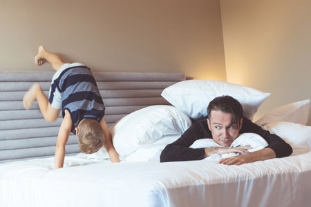 Father covered with pillow lying down on the bed while son is being mischief and climbing on the bed wall.の写真素材