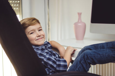 Smiling kid using computer at home and relaxing in chair with feet up on table while looking at camera.の写真素材