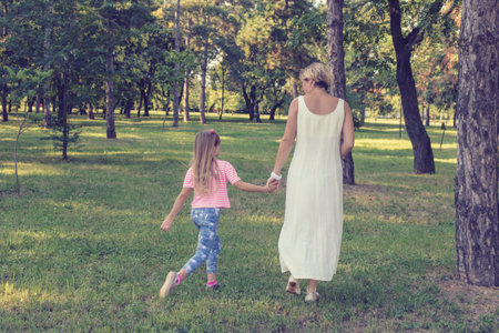 Back view of mother and daughter holding hands and walking through park.の写真素材