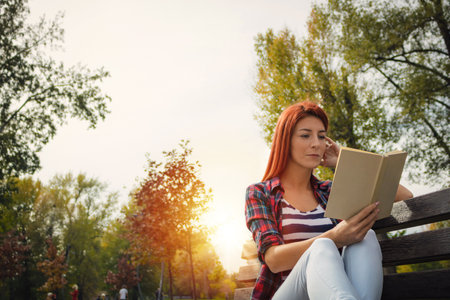 Beautiful red-haired girl reading a novel in the park. Pensive woman sitting on park bench and reading a book.の写真素材