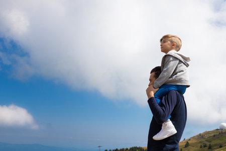 Father carrying his son on shoulders while standing on top of the mountain and looking at view against the sky.の写真素材