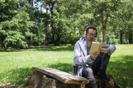 Mid adult man reading book and enjoying in good music over headphones in park during spring day.の写真素材