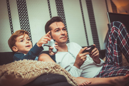 Small kid and his dad playing video games while relaxing on the sofa in the living room.