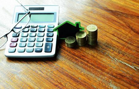 Mortgage loans concept with house sign and coins stack and calculator and eyeglasses on wooden table backgroundsの写真素材