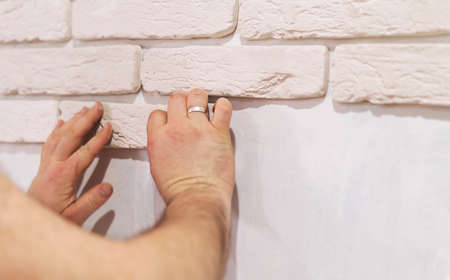 Men's hands glue decorative white bricks to the wall. A professional builder lays decorative gypsum...