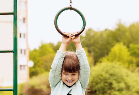 A happy little girl is holding onto an old iron gymnastic ring. A smiling, cheerful little girl is hanging on a gymnastic ring, playing on the playground. Summer active sports leisure for children.の写真素材