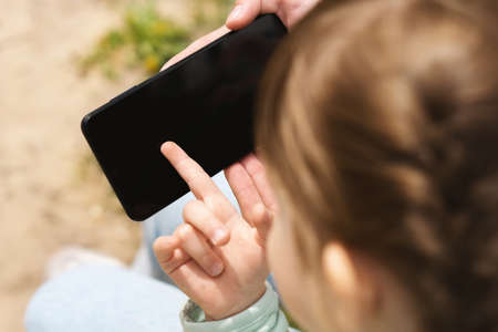 A little girl points her finger at a black blank mobile phone screen. The child uses a smartphone. A cell phone in the hands of a child on the street. The concept of teaching children to use gadgets.の写真素材