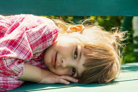 Portrait of a pretty little girl lying on a park bench. Close-up of a cheerful little girl lying on a bench in the open air and looking at the camera with a smile.The child is resting in the fresh airの写真素材