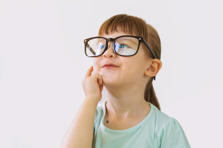 A thoughtful little girl with glasses. Cute thoughtful blonde girl standing on a white background. A beautiful child playing the thinker. The little girl is up to something. Solutiの写真素材