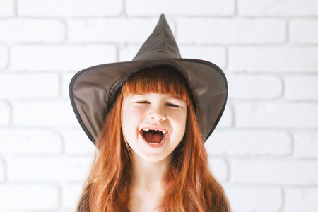 A cheerful little girl with long red hair is smiling in a witch costume for Halloween.A cute little girl with a toothless smile in a witch hat.The red-haired witch.Halloween for kids, a place for textの写真素材