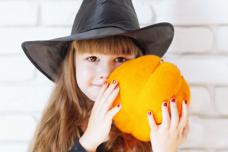 A little cheerful girl holds an orange pumpkin. A child in a black hat is getting ready for...
