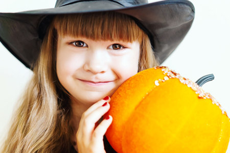 Funny Little Girl In A Halloween Witch Costume With A Pumpkin looks at the camera. A beautiful baby with a pumpkin celebrates Halloween. A pretty little witch with a pumpkin in her hands close-up.の写真素材