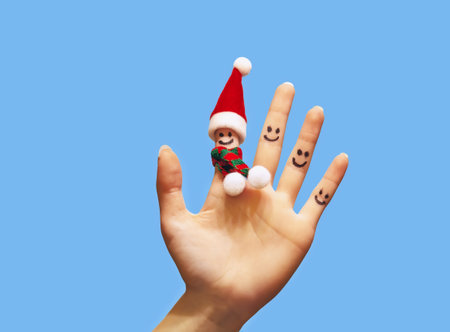 A woman\'s hand with a red Christmas hat on her finger and a scarf with buboes on a blue background. Close-up of a tiny Santa Claus hat on his finger with a smiley face.の写真素材