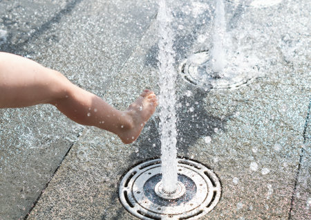 The bare foot of a child in the spray of the city fountain. A child's foot in the stream of a fountain spouting from the ground. Water splashes from under the child's feet. Children's water games.の写真素材