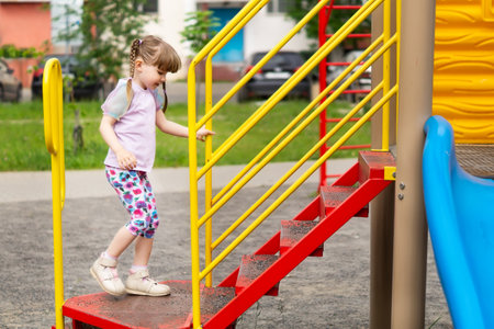 Cute little girl climbs the steps of the slide on the playground, side view. A happy pretty little girl with long pigtails is playing on an outdoor playground. Active children's outdoor gamesの写真素材