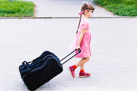 Happy little girl with a big suitcase, ready to travel. A little girl pulls a suitcase on wheels along a city street. An independent 5-year-old girl walks with a travel bag on wheels. Vacation, travelの写真素材