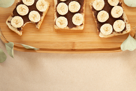 Three banana white bread toasts smeared with chocolate butter that lie on a cutting board with a sprig of leaves on craft paper background. top view with area for textの写真素材