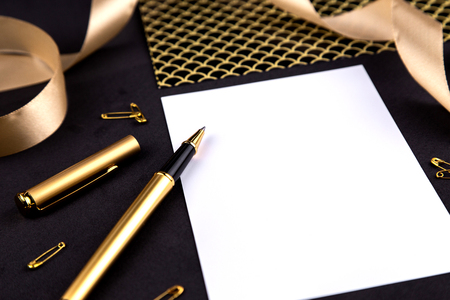 Gold pen, ribbon, paper clips and stationery on a black background with a white sheet of paper.の写真素材