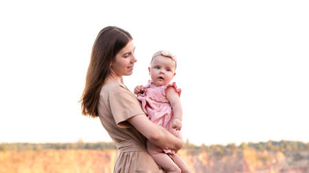 Happy young mother holds her young baby daughter in her arms outdoors. Mom smiles and looks at her daughter.の写真素材