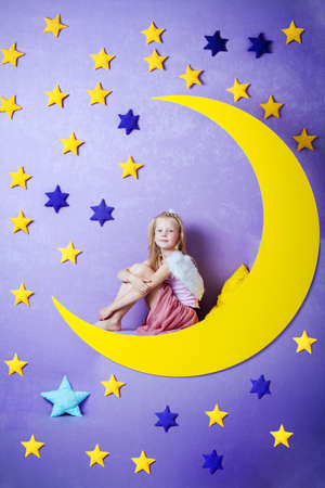 cute little girl with angel wings sitting on a big moonの写真素材