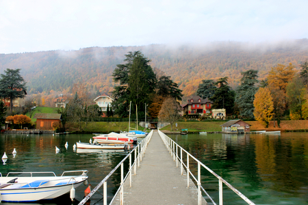 A wooden pier extends into a crystal clear alpine lake, Lake Annecy, where fishing boats dock in the city of Annecy in Autumn, France.の写真素材