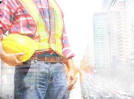 dubble exprosere of engineer in safety suit holding yellow helmet with blur city scape background , construction concept.の写真素材