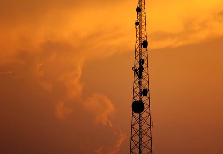 the telacommunication pole with sunset sky background. the silhouette electric workers working on high ground with safety uniform.の写真素材