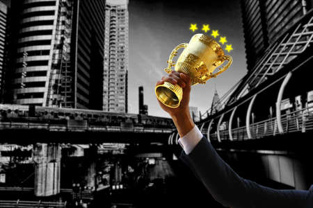 business hand in suit holding golden trophy with blur city scape in gray . illustation stars on golden trophyの写真素材
