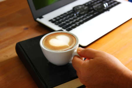 hand holding morning coffee in relax time . heart shape surface on white coffee cup on black book with  office suppliesの写真素材