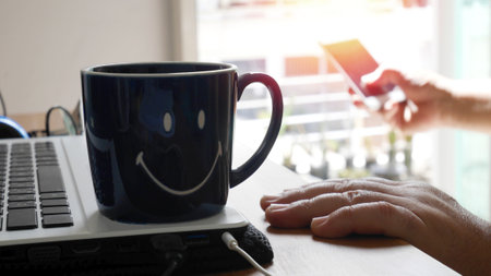 ceramic coffee cup on laptop with flare on blur hand using mobile phone . man using smartphone work at home with coffee cupの写真素材