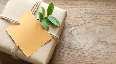 empty card on gift box wrapped in recycled paper with rope and leaf on wood background with copy...