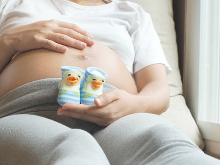 Pregnant woman holding small shoes for the unborn baby in front of the belly.