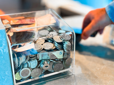 Transparent donation box filled with various coins and banknotes on countertop with hand holding credit card in background. Close up shot for financial aid and charity concept.の写真素材