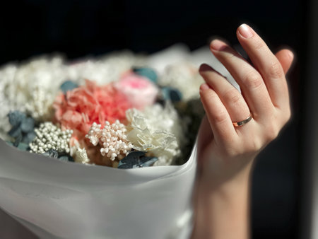 Close up of woman hand wearing gold ring gently touching bouquet of flowers. Delicate mix of white, pink, and blue blooms wrapped in soft paper. Wedding concept.の写真素材