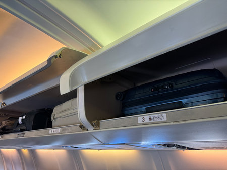 Overhead storage compartment inside airplane cabin filled with various carry on luggage, including black and white suitcases, backpacks, bags, with compartment doors open, visible seat row numbers.の写真素材