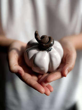 Close up of hands gently holding small white ceramic pumpkin with twisted brown stem against blurred background. Minimalistic and symbolic autumn or Halloween decor and seasonal.の写真素材