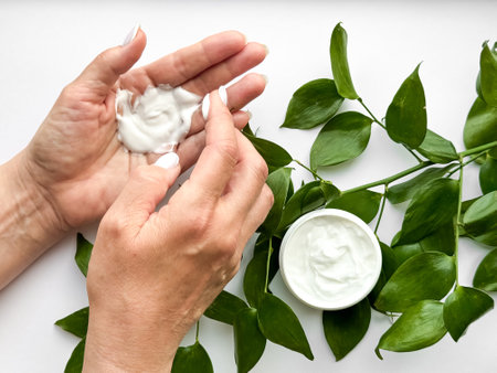 Close up of middle age woman hands applying hand cream with green leaves on white background. Skincare and natural beauty concept. For healthcare, wellness, and organic product poster.の写真素材