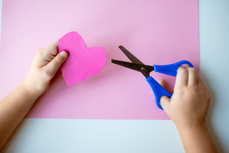Childs hands cutting pink paper heart with scissors on pink backgroundの写真素材