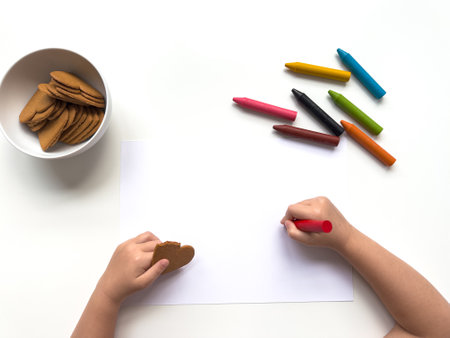 Child drawing with crayons and eating cookies on a white surfaceの写真素材
