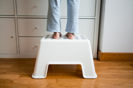 Child standing on white step stool reaching high shelf in home setting