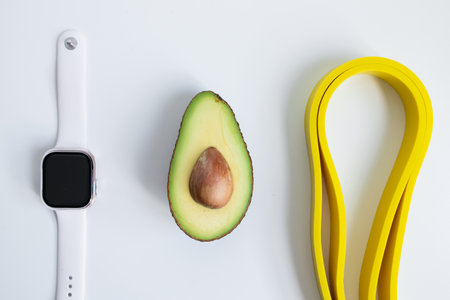 Minimalist arrangement with smartwatch, avocado, and yellow resistance bands on white background