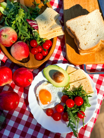Delicious picnic spread with avocado, cheese, eggs, apples, tomatoes, green leaves, and bread on checkered clothの写真素材