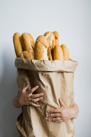 Child holding large paper bag filled with french baguettes against plain background
