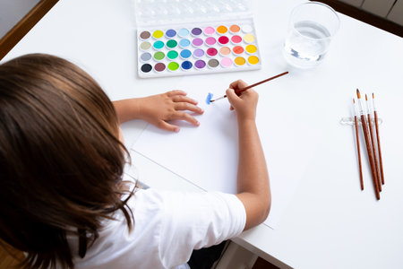 Young caucasian child painting with watercolors on white paper at a tableの写真素材