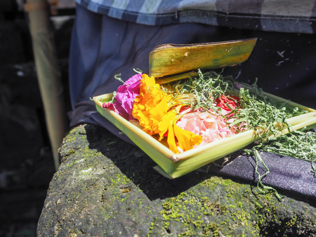 Balinese offering basket made out of palm leaves and filled with flowers, at the base of a stone statue, Besakih temple, Bali, Indonesiaの写真素材