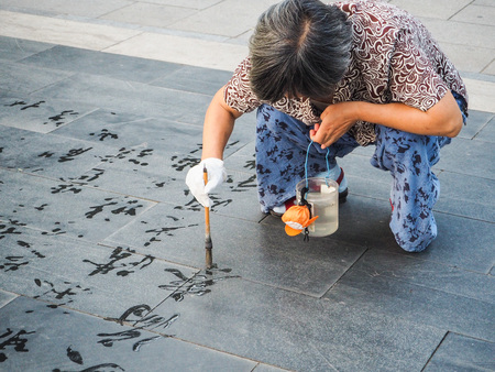 Older woman practicing water calligraphy but due to the heat the Chinese characters disappear immediately and are not readable, Beijing, Chinaのeditorial素材