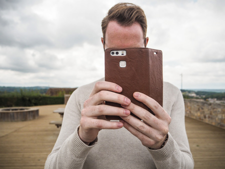 Young man with brown hair standing outdoor and taking a picture with his smartphone which has a...
