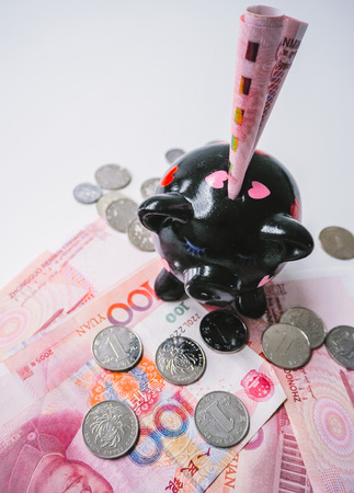 Black piggy bank standing on Chinese 100 rmb banknotes and coins on a white wooden background. The pig symbols fortune in the Chinese zodiac.の写真素材